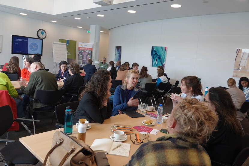 Aan zes langwerpige tafels zitten deelnemers van de townhall-sessie. Zij zijn per tafel met elkaar in gesprek over een workshop.