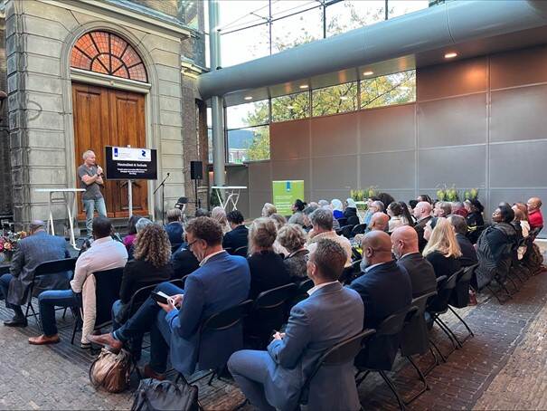 In de glazen zaal van de Liberaal Joodse Synagoge in Den Haag kijkt het publiek naar een man die aan het spreken is. De man heeft een microfoon vast en naast hem staat een televisiescherm met een presentatie.